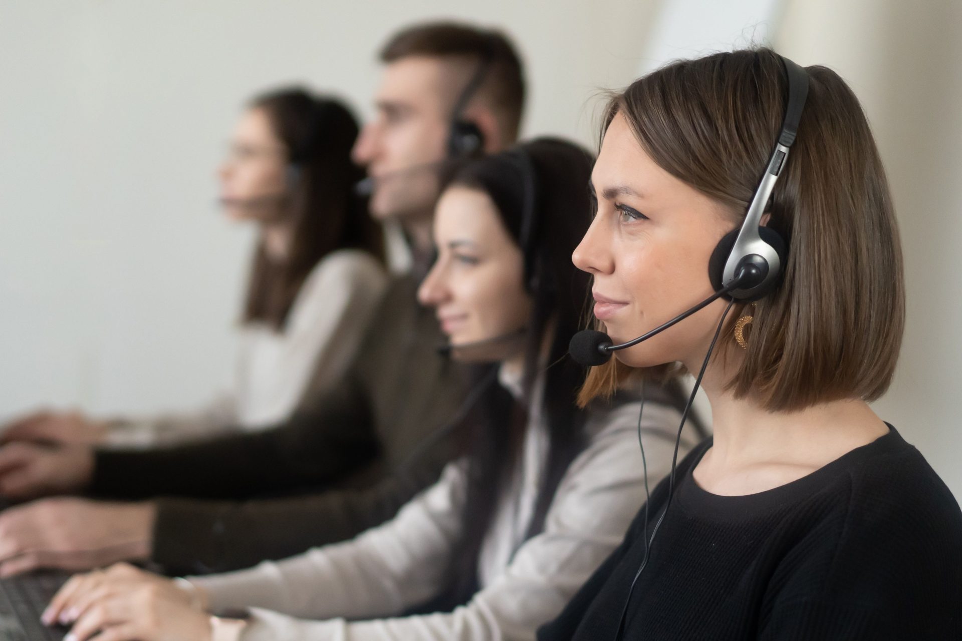 Seitenansicht junger Kollegen in Freizeitkleidung, die mit Headsets (inklusive Mikrofon) an einem hellen Arbeitsplatz sitzen und am Laptop surfen.