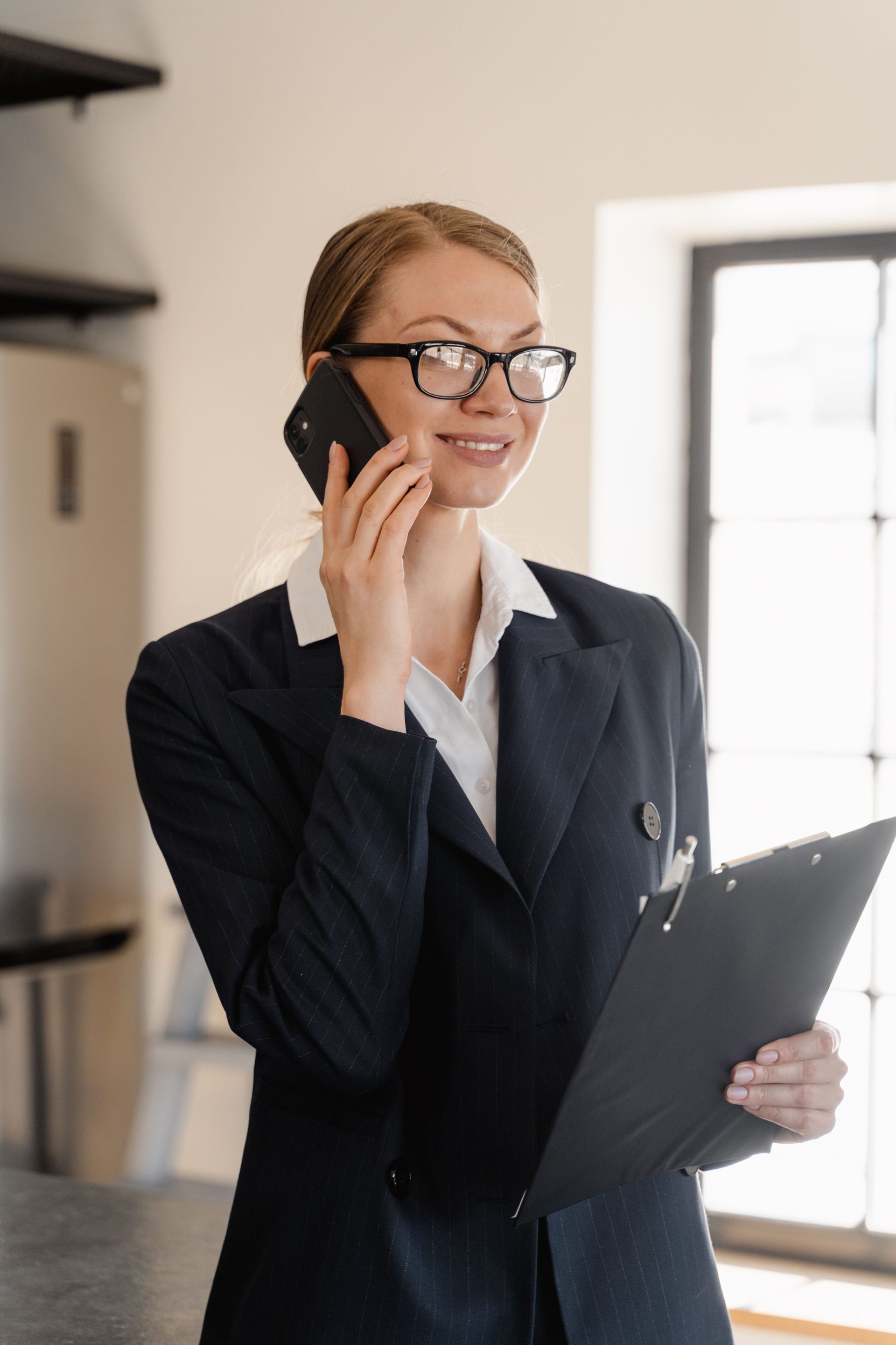Lächelnde Geschäftsfrau in Business-Kleidung telefoniert und hält ein Klemmbrett in einem hellen Büro.